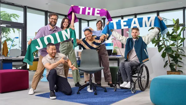 A diverse group is holding scarves with the inscription “Join the Team” in the air and is pointing to an empty office chair.